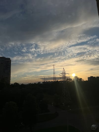 This is a vertical, outdoor, full shot of a sunset scene over a suburban area, likely Shushary, Russia. The sky is filled with dramatic clouds, some dark and heavy on the left, transitioning to wispy, elongated clouds illuminated by the setting sun on the right. The sun itself is a bright orb, casting strong rays of light that create lens flare effects.

Prominent in the midground are two large electrical pylons, silhouetted against the bright sky. Their lattice structures are clearly visible. Beyond the pylons, a dense line of dark trees forms a treeline separating the sky from the ground. On the far right, a road curves through the scene, with streetlights and a crosswalk visible. A parked car with its taillights illuminated is also present on the road.

In the foreground, on the bottom left, a portion of a tall, multi-story apartment building is visible, also in silhouette. On the top right, another apartment building can be seen.

The overall mood is serene and slightly dramatic, with the vibrant sunset contrasting with the dark, silhouetted elements of the urban landscape. The time of day is clearly sunset due to the low angle of the sun and the characteristic lighting. The weather appears to be partly cloudy. No discernible text is present in the image.
