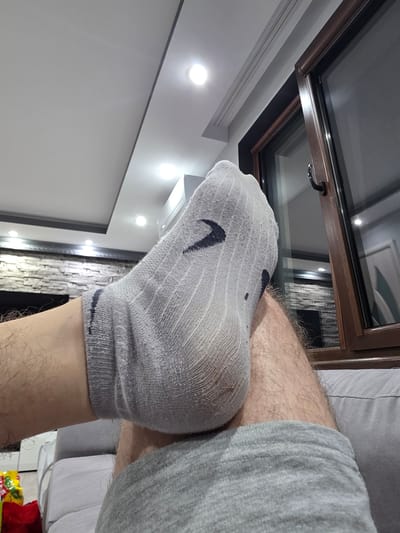The image depicts an indoor scene in Yagodovo, Bulgaria, viewed from a low angle, focusing on a person's elevated lower legs and the surrounding environment. The primary subject is a hairy leg, wearing a light grey, ribbed athletic sock with a dark grey or black swoosh-like logo visible on the top of the foot. The sock exhibits signs of wear and dirt, particularly around the heel. The foot is elevated, suggesting a resting posture, with the leg possibly resting on the person's other leg or a piece of furniture. The person is wearing light grey athletic-style shorts, visible on the lower right.
The background reveals a contemporary living space. To the right, a large, dark-framed window, possibly double-glazed, extends upwards, showing a handle and reflecting elements of the room's interior, including a white door with a distinct curvilinear panel design and a portion of the textured wall. Above the window, a white ceiling features a recessed section and multiple bright, circular LED spotlights. To the left and center-rear, a feature wall constructed from light-colored, stacked stone or brick veneer is visible, with a dark, rectangular recess below, possibly indicating a fireplace. Additional ceiling lights are scattered across the white ceiling. A grey upholstered sofa or couch is partially visible in the lower foreground and lower right, indicated by its material and stitching. In the extreme lower-left corner, a red bag and a colorful, crinkled package, possibly containing snacks, are partially visible on what appears to be the floor or a low surface. The overall lighting is artificial and bright.