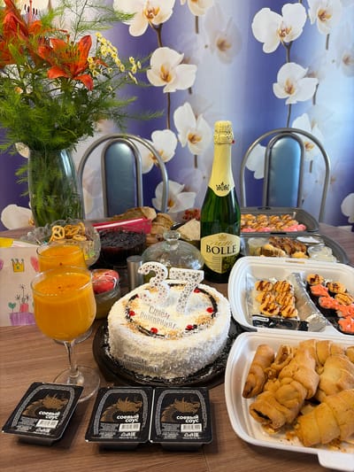 The image depicts a table set for a birthday celebration, with a cake, champagne, sushi, drinks, and pastries. The background features a wall with a floral pattern, likely orchids, in shades of blue and white. There are no people visible in the image. The arrangement suggests a festive occasion with various food and beverage items. The presence of specific items like sushi and a decorated cake indicates a celebratory meal. The overall setting appears to be indoors, possibly a dining area or a room prepared for a party.