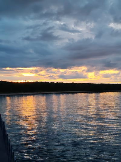 The image captures a sunset over a body of water, possibly a lake or a wide river, in the village of Shcheleiki, Russia. A dark, treed shoreline separates the water from the sky. The sky is filled with dramatic, heavy clouds, tinged with orange and yellow where the sun is setting below the horizon. The setting sun casts a bright, golden reflection on the rippling surface of the water. In the foreground, the edge of a dark structure, likely a pier or boardwalk, is visible on the left side of the frame. There are no people or animals visible in the scene. The overall atmosphere is serene and perhaps a little moody due to the dark clouds.