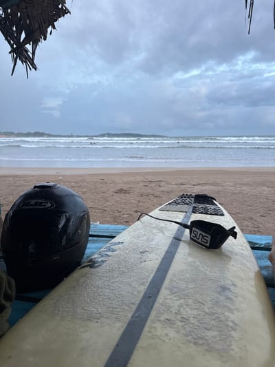 The image depicts a close-up of a surfboard and a black helmet resting on a blue surface, likely a bench or a table, on a beach. The scene is set at Weligama, Sri Lanka. The ocean with gentle waves is visible in the background, along with a sandy beach. The sky is overcast and appears to be a cloudy, possibly rainy, day.
The surfboard is light-colored with a dark stripe running down its length. A black leash with a white logo that reads "SUHS" is attached to the tail of the surfboard. The helmet, also black, is positioned in front of the surfboard and appears to be wet, suggesting recent rain. The textured surface of the surfboard, possibly from wax or wear, is visible. To the left of the frame, a portion of a thatched structure is present, hinting at a beachside shack or shelter.
The activity suggested by the presence of a surfboard and helmet is likely related to surfing or a similar water sport, although no people are actively engaged in these activities in the frame. The weather conditions suggest a challenging or overcast day for outdoor activities. The overall mood is somewhat muted due to the weather, but the setting conveys a sense of a coastal recreational area.