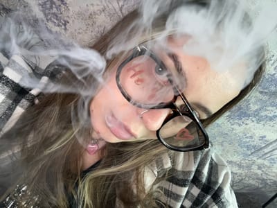 The image shows a person lying down, exhaling a cloud of vapor. The person is wearing glasses, which reflect an image, and has blonde hair and piercings in their nose and ear. They are lying on a surface with a patterned fabric. The location, Novopokrovskaya, Russia, is not visually discernible from the image.