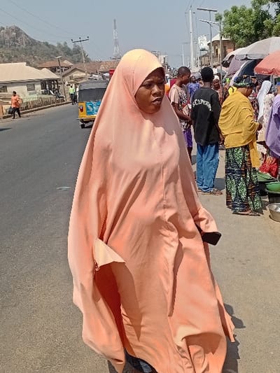 A woman in an orange hijab stands on a roadside, facing the camera. Behind her, people and stalls suggest a market scene. The background shows a landscape with buildings and a hill. The woman's expression is neutral, and the hijab covers her head and body, with a section on the ground. The setting appears to be a bustling outdoor environment with varied activities.