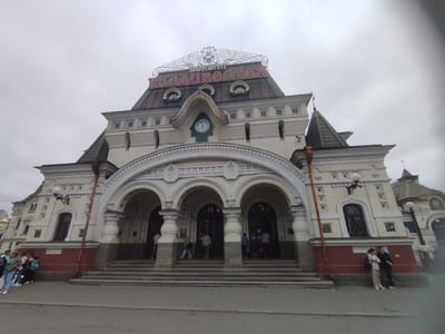 The image shows the exterior of Vladivostok Railway Station, a historic building in Vladivostok, Russia. The station is an ornate, cream-colored building with red accents and a prominent clock face. Above the main entrance, large red letters spell out "ВЛАДИВОСТОК" (Vladivostok) on the roof, indicating the station's name. The sky is overcast, suggesting a cloudy day. Several people are visible on the steps and sidewalk in front of the station. Some are standing in groups, while others are walking or entering the building. The architecture features multiple arches, columns, and decorative elements, typical of late 19th-century Russian architecture. The overall scene is one of a busy public transportation hub on a somewhat gloomy day.