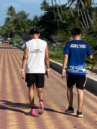 Two individuals, viewed from behind, are walking on a paved path with a patterned surface of yellow and red stripes. The person on the left wears a white sleeveless shirt with blue text that reads "HYBRID TRAINING CLUB" and black shorts. They have tattoos on their left arm and wear a black baseball cap, a white wristband on their left wrist, and pink and grey athletic shoes. The person on the right wears a blue t-shirt with white text that reads "PHAN THIET RUN & FUN" and a white and blue graphic design along the bottom. They also wear black shorts and black athletic shoes. Both individuals have white wristbands on their right wrists.
The background features a vibrant tropical environment. Lush green palm trees and other foliage line the right side of the path, with tall palm trees in the distance. Behind the foliage, parts of buildings and structures, some appearing weathered or under construction, are visible. Boats are also present near the shoreline, suggesting a coastal or near-water location. The path appears to be an elevated walkway or promenade, with a low concrete barrier on the right side separating it from the background. The lighting suggests it is daytime, with clear skies and shadows cast on the path. The location is Mũi Né, Vietnam, a coastal town known for its beaches and watersports.