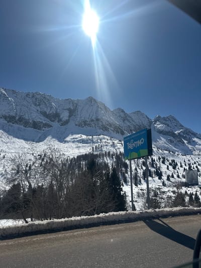 The image presents a snow-covered alpine landscape in Vermiglio, Italy, under a clear blue sky with a prominent, bright sun generating a starburst lens flare in the upper center. Majestic, snow-capped mountains with visible rock faces and rugged ridges dominate the background. Below the peaks, snowy slopes are interspersed with evergreen coniferous trees and leafless deciduous trees. In the midground, a large billboard sign is visible, displaying the word "TRENTINO" in white letters on a blue and green background, with smaller text "PREMIERE ALPENREGION GONDEL" above it. The sign is supported by two dark poles. Further on the right, in the background, a tall, multi-story building of a grayish tone stands against the snowy mountain. The foreground reveals a paved road surface, with a substantial snow berm lining its right side. Shadows are cast onto the road, suggesting the perspective is from a vehicle. No individuals or specific actions are depicted.