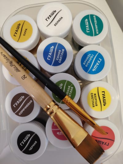 This is a close-up, top-down shot of a set of gouache paints in a clear plastic container, along with two paint brushes. The container holds multiple small, round tubs of paint with white lids, each featuring a colored label indicating the paint color and name in Russian.

The visible paint colors and their Russian labels are:
*   Yellow: "ГУАШЬ ОХРА" (Gouache Ochre)
*   White: "ГУАШЬ БЕЛИЛА" (Gouache White)
*   Teal/Green: "ГУАШЬ ИЗУМРУДНАЯ" (Gouache Emerald)
*   Blue: "ГУАШЬ СИНЯЯ СВЕТЛАЯ" (Gouache Blue Light)
*   Dark Blue: "ГУАШЬ ГОЛУБАЯ ТЕМНАЯ" (Gouache Blue Dark)
*   Green: "ГУАШЬ ЗЕЛЕНАЯ СВЕТЛАЯ" (Gouache Green Light)
*   Yellow: "ГУАШЬ ЖЕЛТАЯ СВЕТЛАЯ" (Gouache Yellow Light)
*   Brown: "ГУАШЬ КОРИЧНЕВАЯ" (Gouache Brown)
*   Black: "ГУАШЬ ЧЕРНАЯ" (Gouache Black)
*   A partially visible pink/red label reads "ГУАШЬ".

Two paint brushes are positioned diagonally across the paints. One is a flatter brush with a wide, golden ferrule and a wooden handle inscribed with "Mr.Painter" and "20". The other is a thinner brush with a black handle and a golden ferrule.

The lighting is bright, suggesting it was taken indoors under artificial light or near a window during the day. The setting appears to be a home or art studio in Balashikha, Russia, given the context. The image focuses on the art supplies, implying an artistic activity. There are no people visible.