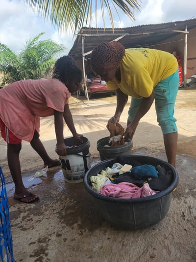 Two women are engaged in washing clothes outdoors in what appears to be Madalla, Nigeria. One woman, wearing a yellow shirt and teal capri pants with a net cap on her head, is bent over a gray bucket, washing clothes. The other woman, in a pink top and red shorts, is similarly positioned over a white bucket with black lettering. A large, dark gray basin filled with clothes sits nearby. The ground is wet, and the surrounding environment includes a corrugated roof, palm trees, and a glimpse of a red vehicle.