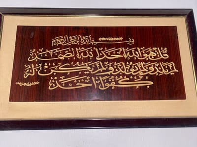 This media file is a close-up, eye-level shot of a framed piece of Islamic calligraphy. The artwork is mounted on a dark, polished wood plaque and framed with a dark brown border. The calligraphy itself is in a golden, raised font, set against the rich, dark wood. The inscription appears to be from the Quran, with the top line including "بِسْمِ ٱللَّٰهِ ٱلرَّحْمَٰنِ ٱلرَّحِيمِ" (In the name of Allah, the Most Gracious, the Most Merciful). Below this, larger Arabic script is visible, followed by a smaller inscription in the bottom left corner which reads "صدق الله العظيم" (Allah the Almighty has spoken the truth). The overall impression is one of religious devotion and artistic craftsmanship. The lighting is even, suggesting indoor display. No specific time of day, weather, or location cues are visible beyond the context of Fagge A, Nigeria.