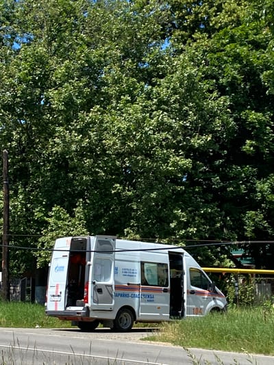 A white van with emergency markings is parked on the side of a road in what appears to be a suburban or rural area of Zapadny, Russia. The van's rear doors and the side passenger door are open, revealing equipment and personnel inside. The van has the logo of "GAZPROM" and what appears to be emergency service markings in Russian. A large, leafy tree dominates the background, with a wooden utility pole and wires in the foreground. The weather is sunny, and the vegetation is green and lush, suggesting a warm season.