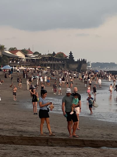 The image captures a crowded beach scene in Canggu, Indonesia, with numerous people milling along the shore. The sky is overcast, contributing to a muted color palette. The beach is wet, reflecting the figures and structures. A temple complex with multi-tiered structures is visible in the background. Close to the viewer, people are standing, walking, and looking towards the ocean. To the far right, there is what appears to be a retaining wall.