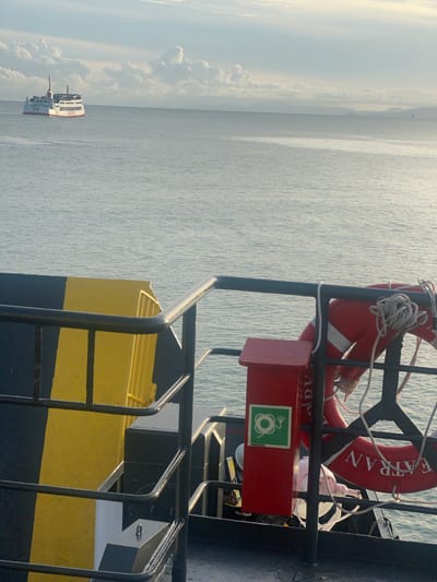This is a vertical, medium-shot photograph taken from a boat. The scene is the sea with a cloudy sky. In the foreground, on the right side of the image, a red fire extinguisher box with a green symbol of a hose and coiled rope is mounted on a metal structure. Above the box, a red life preserver with a white stripe and the word "SETRAN" visible is suspended. The water is calm and appears to be a light blue-grey. In the distance, on the left side of the image, a white ferry with red trim is visible on the water. The sky is bright, suggesting it is daytime, possibly early morning or late afternoon, with diffused sunlight creating a soft, atmospheric light. The overall mood is serene and suggests a journey at sea. There is no clear indication of people in the image, though their presence on the ferry is implied. The location context is unknown, Thailand, as per the prompt, but the scene is typical of ferry travel in many coastal regions.