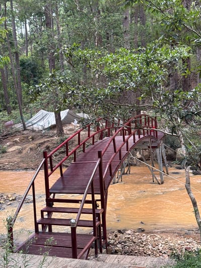 The image shows a metal pedestrian bridge spanning a murky, brown river or stream. The bridge is painted red and has a curved design with steps leading up to it from one side. The surrounding environment is lush with greenery, including trees with dense foliage. In the background, there are structures that appear to be greenhouses or covered agricultural areas. The setting is likely a rural or semi-rural area, possibly near the city of Lang Biang - Đà Lạt, Vietnam, known for its agriculture and natural landscapes. The absence of people suggests a tranquil and possibly remote location.