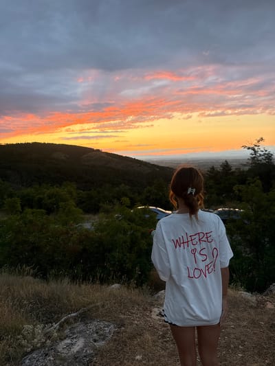 A young woman stands with her back to the camera, looking out at a scenic sunset over a valley. She is wearing a white t-shirt with red lettering that says "WHERE IS LOVE?". The t-shirt is partially obscured by her hair, which is pulled back in a bun with a hair clip. She is also wearing a black and white striped skirt. The woman is standing on a dirt path, with dry grass and rocks around her. In the background, there are trees and hills, and the sky is filled with colorful clouds. The location is the area surrounding the city of Markovo, Bulgaria.