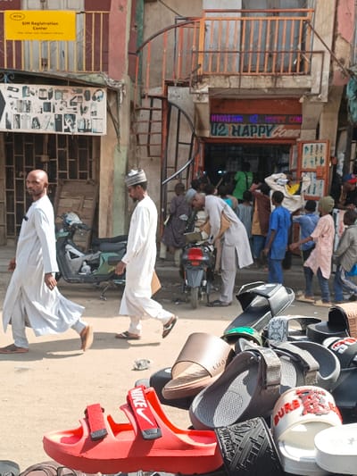 The image depicts a busy commercial street scene in Fagge C, Nigeria, under bright daylight. In the immediate foreground, a street vendor's display of various sandals and slides is visible, featuring items in red (with "NIKE" branding), white (partially "BURBERRY"), black (partially "LOUIS VUITTON"), and brown ("LUODONG").
In the midground, two adult males, both dressed in light-colored traditional tunics and sandals, walk from left to right. The lead male, bald, glances towards the viewer, while the second male, wearing a skullcap, looks downward. Behind them, a green and black scooter is parked. Further right, a crowd of people, including several children and adults in varied attire, are gathered around the entrance of a shop. One adult male, wearing a light-colored tunic, is bent over near a parked black motorcycle.
The background features multi-story commercial buildings. On the left, a building displays a prominent yellow "MTN SIM Registration Center" sign. Below it, a vertical display board showcases mobile phone accessories. On the right, a shop named "U2 HAPPY Comm" (likely a telecommunications store) has a neon blue and green sign and is brightly lit internally, with customers at its entrance. Above this shop, an upper floor features an orange-brown balcony railing and an external black spiral staircase. The ground surface is unpaved, appearing dusty.