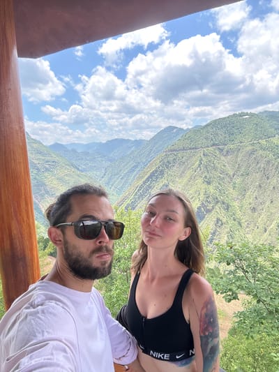 Two people are in a selfie photograph with a scenic mountain background. The woman is fair-skinned, wearing a black sports bra with "NIKE" visible, and has a tattoo on her upper arm. The man has a beard, sunglasses, and is wearing a white t-shirt. They appear to be on a balcony or platform with a wooden railing. The background features a steep, verdant mountain landscape under a partly cloudy sky. The location is Lũng Cú Commune, Vietnam, indicated by the mountainous landscape typical of the region.