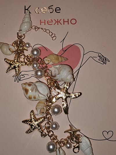 The image displays a close-up of a collection of charms, including seashells, pearls, and starfish, attached to a gold-toned chain. These charms are arranged against a light pink background with some black line art that suggests a human figure. Text in Russian, "К себе нежно," meaning "To oneself tenderly," is visible at the top of the background. The overall presentation suggests a decorative item, possibly jewelry or a charm bracelet. There is no visible indication of the city of Tyrnyauz, Russia, in the image.