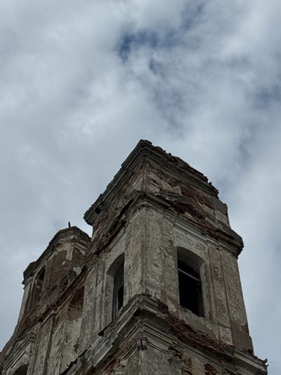 The image shows a close-up, low-angle view of the dilapidated upper sections of a stone building, likely a church or similar structure, against a cloudy sky. The building appears to be abandoned, with visible damage to its facade, including crumbling plaster and exposed brickwork. Arched window openings are dark and appear empty. The architecture suggests historical significance, with decorative molding and cornices evident despite the decay. The scene is set in the Княжыцкі сельскі Савет, Belarus, a rural administrative division. The overall atmosphere is one of neglect and the passage of time.