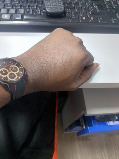 The media file shows a close-up view of a person's right hand and wrist, positioned over a keyboard and next to a white surface and a blue drawer. The person is wearing a dark wristwatch with a black band and a circular face featuring gold-colored hands and sub-dials, and some red accents. The skin tone of the hand suggests a person of color, and a fingernail is visible on one of the fingers.
The background includes a black keyboard with visible keys, a white vertical surface that could be a desk or cabinet, and a partially opened blue drawer with metal slides. To the right of the white surface, a wooden surface is visible. The lighting suggests an indoor setting, likely during the daytime, but no specific time or weather conditions can be determined. The overall scene appears to be an office or study environment.
No specific activity or event is clearly depicted, other than the hand resting on or near the keyboard. The image is focused on the hand and wrist, with the surrounding objects providing context. There is no discernible text in the image, except for a faint logo or text on the watch face that is not clearly legible. The setting is identified as Fagge A, Nigeria.