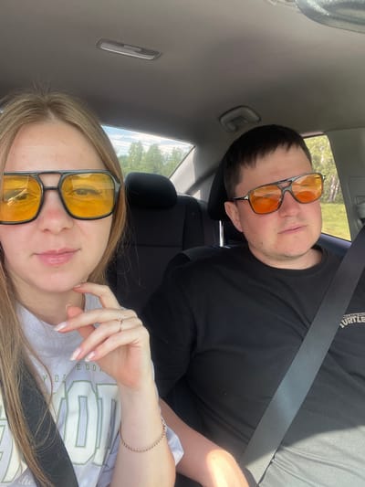 In the city of Brodokalmak, Russia, two individuals, a woman and a man, are seated inside a vehicle, likely a car. They are both wearing distinctive sunglasses with orange lenses. The woman, positioned on the left, has blonde hair and is looking towards the camera, touching her chin with her right hand, revealing a ring on her ring finger and a bracelet on her wrist. She is wearing a white t-shirt with light green lettering. The man, on the right, has short dark hair and is also looking towards the camera. He is wearing a black t-shirt with a graphic print of what appears to be "Teenage Mutant Ninja Turtles" on the right side of his chest. Both individuals have their seatbelts fastened. The interior of the car is visible, including the headrests and the ceiling. Through the windows, a bright, sunlit outdoor environment with trees and foliage can be seen, suggesting it is daytime and likely a rural or suburban setting.