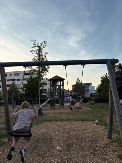 This media file captures a playful scene at a playground in Bönnigheim, Germany. Two children are enjoying swings under a bright, slightly cloudy sky, suggesting it's likely late afternoon or early evening. The playground features a wooden swing set with chains and black seats. A young girl with blonde hair, wearing a light-colored top and a skirt, is actively swinging forward. Behind her, another child, also with blonde hair, wearing a white t-shirt and khaki shorts, is on a separate swing, moving backward. The ground beneath the swings is covered with wood chips for safety.

In the background, a large, multi-level wooden playground structure with a tower and slide is visible. Beyond that, modern white apartment buildings and lush green trees line the horizon, providing a residential context to the playground. A soccer ball rests on the wood chips near the other swing set. A single pink shoe is also visible on the ground. The overall atmosphere appears joyful and relaxed, typical of children engaged in outdoor play. The lighting suggests a natural, possibly golden hour light, creating a warm ambiance. No visible text is present in the image.