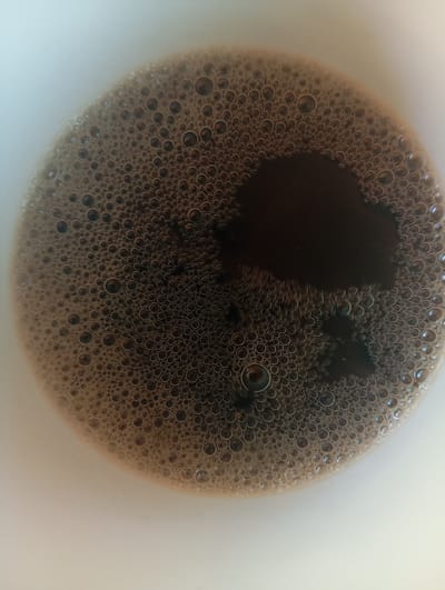 This is a close-up, top-down view of the surface of coffee in a white mug. The coffee has a thick layer of small, dark brown bubbles covering most of the surface. There is a darker, more liquid area in the upper right quadrant, suggesting that the coffee is not uniformly mixed. The mug appears to be made of white ceramic. No specific location in Orenburg, Russia, can be determined from this image.