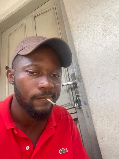 A man wearing a red polo shirt and a brown baseball cap is seen from the chest up, smoking. He has a beard and is looking towards the camera. Behind him is a light-colored door and a textured wall with a door handle and keys. The scene appears to be outdoors, likely in front of a building.