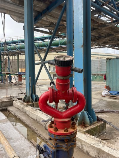 A fire hydrant is centrally located in a semi-industrial outdoor setting. The hydrant is red with a black nozzle and is connected to a blue valve at its base. It is positioned on a concrete platform surrounded by water and debris. In the background, there are large blue metal support beams, industrial pipes, and a portion of a shipping container. The sky is overcast. No people are visible in the image. The location is described as Unknown, Indonesia.