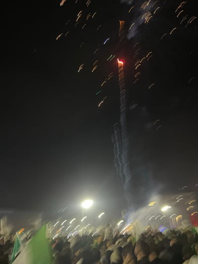 The image shows a nighttime scene with streaks of light, suggesting movement. On the left, a reddish point of light is surrounded by yellow streaks. A wisp of smoke trails away from it. On the right, a blurry illuminated area is visible, marked by vertical streaks of white and other colors, possibly indicating a crowd. The background is predominantly dark, and the overall impression is one of an event or display occurring at night.