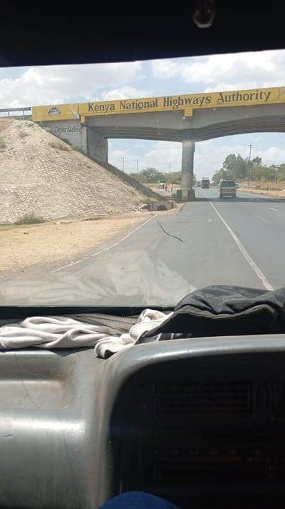 The photo is taken from inside a vehicle, looking out through the windshield. The view is of a highway with a bridge overhead, adorned with "Kenya National Highways Authority" in bold letters. Cars and trucks are visible on the road. To the left of the road, there's a hillside with what appears to be rock or concrete reinforcement. Parts of the dashboard and some clothing are visible in the foreground, indicating the interior of the vehicle.