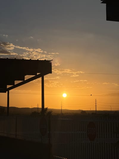 A low-angle, vertical, full shot captures a sunset in Sunland Park, USA. The sun, a bright orange orb, is positioned in the center of the frame, casting a warm glow across the sky. Wispy, white clouds are scattered across the upper portion of the sky, contrasting with the deeper blue hues. A silhouetted structure, possibly a covered patio or awning, juts in from the left side of the image, supported by dark vertical posts. In the foreground, a white slatted fence runs horizontally across the lower third of the image. Two octagonal signs are visible on the fence; one, partially obscured, appears to have a white circle with a red border and a line through it, possibly indicating "Do Not Enter." The other sign, more clearly visible on the right, is a red stop sign. Beyond the fence, a dark, undulating horizon line suggests hills or mountains. Several power lines and utility poles are visible, stretching across the landscape. A small bird is perched on a wire in the middle ground, adding a touch of life to the scene. The overall atmosphere is serene and peaceful, with the warm tones of the sunset creating a sense of tranquility.