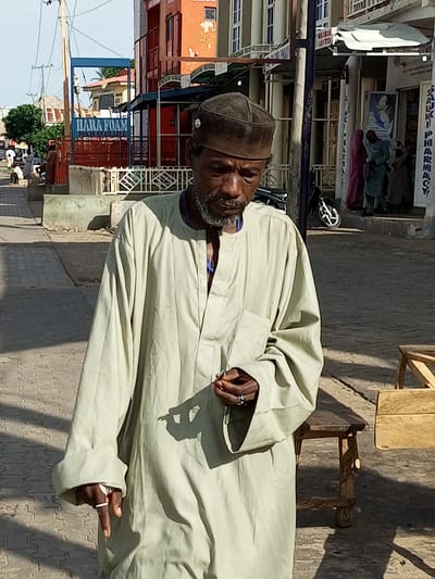 The media file shows a medium shot of an elderly Black man walking on a paved street in what appears to be a market or commercial area in Arawa, Nigeria. He is wearing a light-colored, long-sleeved robe and a dark cap. His face is weathered, and he has a gray beard and mustache. He is holding a small object in his right hand, and his left hand is resting on his chest, holding another object.
The background suggests a bustling street with several buildings. To the left, there's a building with orange and white facades and a blue sign that reads "HARA FOAM". Further down the street, there are shops with awnings and signs, including one that appears to say "SAUKKI PHARMACY". Several other people are visible in the background, some walking and others standing, suggesting it is daytime, likely late morning or early afternoon, given the strong shadows cast on the ground. The weather appears to be sunny and warm. The man's expression is neutral, and the overall scene conveys a sense of everyday life in a Nigerian town. There are also wooden benches and tables visible on the side of the street.