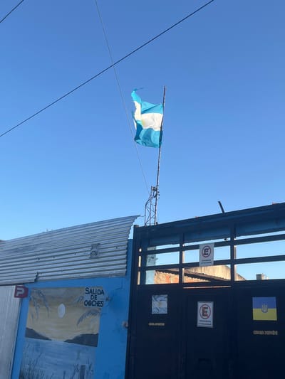 An outdoor daytime scene depicts a street-level view in Partido de Quilmes, Argentina, under a clear blue sky. A tattered Argentine flag, predominantly blue and white with a yellow sun emblem, flies from a metal pole atop a rudimentary metal structure positioned above a dark, likely metal, gate. Two dark utility lines traverse the upper left quadrant of the sky.
To the left of the gate, a section of a corrugated metal wall or roof, silvery-grey and irregularly shaped at its top edge, is visible. Below this, a vibrant blue painted wall features a mural depicting a nocturnal or twilight landscape with water, land, and a large circular moon or sun. White text "SALIDA DE OCHES" is painted above the mural, and a red "P" parking sign is affixed to the blue wall to the left of the text. A small, light-colored object resembling a stylized face is mounted on the corrugated metal above the blue wall.
The dark metal gate, spanning the center-right of the frame, is composed of vertical and horizontal bars. Two white "PROHIBIDO ESTACIONAR" (No Parking) signs, featuring a red circle with a diagonal line over a black 'E', are affixed to the gate at different heights. On the lower right panel of the gate, a small Ukrainian flag sticker is visible. Another square graphic, light-colored and abstract, is present on the lower left panel of the gate. Portions of other structures, including a reddish-brown brick wall and a dark roof, are discernible through the gate's openings in the background. No individuals are present.
