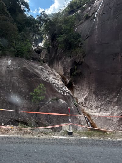 The image depicts a rocky cliff face adjacent to a paved road. The cliff is dark gray and appears to be made of granite, with visible cracks and fissures. Patches of green vegetation, including small trees and bushes, grow out of the rock in several places. A narrow stream of water trickles down the cliff face in a central gully.
Two orange ropes are stretched diagonally across the scene, one appearing to be secured to a metal pole with a black cylindrical object attached. This suggests some form of activity or construction is taking place, possibly related to rock stabilization or a survey. The ropes are taut and extend from the cliff face towards the road.
The road itself is asphalt and shows a white dividing line. The surrounding area is a natural landscape, with lush green trees visible in the upper left portion of the image, and a cloudy blue sky. The lighting suggests it is daytime, with diffused sunlight. There is no clear indication of the specific time of day or weather conditions beyond the clouds.
The location is identified as Nam Khanh Vinh, Vietnam. There is no visible text in the image. The scene conveys a sense of rugged natural beauty combined with human intervention.