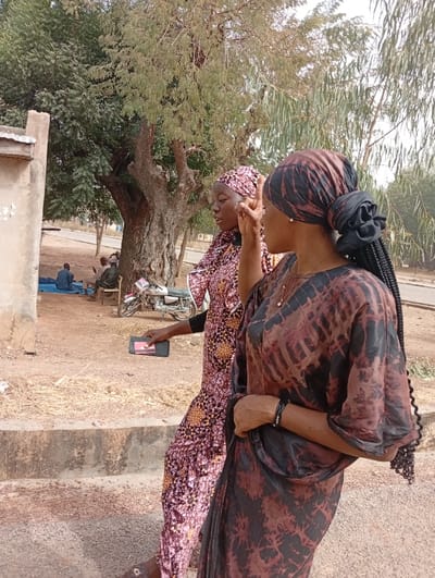 Two women are walking outside. The woman in the foreground is facing right, wearing a brown and black patterned head wrap and dress. She has a braid down her back. The other woman is behind her, wearing a pink and orange patterned head wrap and dress. She is holding a black smartphone in her hand and pointing her finger upward. In the background is a tree, a motorcycle, and two men sitting on the ground. There is a building to the left and a road to the right.