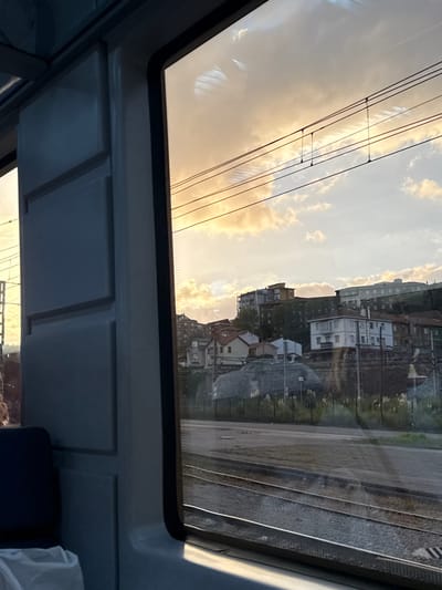 The image captures a view from inside a train or tram in Santander, Spain, depicting an urban landscape under a sky illuminated by golden light, indicating either sunrise or sunset. The interior of the vehicle, visible on the left, consists of light grey, modular paneling and a dark-colored seat with a crumpled white object on it. Reflections of interior ceiling vents are visible on the windowpane.
Outside the window, multiple horizontal overhead electrical lines for the train are stretched across the upper sky. Below these lines, the terrain rises, featuring a dense arrangement of residential buildings on a hill. These structures vary from smaller houses with reddish-brown tiled roofs to larger, multi-story apartment blocks. At the base of the hill, a large, dark grey, irregular structure, possibly a rock formation or concrete barrier, is partially covered by a fine net or tarp. Two parallel railroad tracks, appearing damp or reflective in sections, run across the lower mid-ground, bordered by sparse, tall grass and weeds. No individuals are visible in the scene.