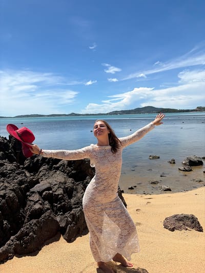 A woman stands on a sandy beach in Baan Bo Phut, Thailand, holding a red hat in her left hand. She is wearing a long, white lace dress and her right arm is extended towards the water. The ocean is calm and blue, with a distant island and green hills visible. The sky is bright blue with scattered white clouds. Dark volcanic rocks are on the left side of the image, and smaller rocks are scattered on the beach and in the water.
