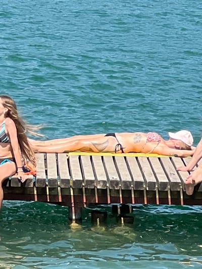 The image depicts a sunny day at a waterside location, possibly a pier or a dock, in Vilnius, Lithuania. In the foreground, a woman with long, light brown hair is sitting on the edge of a wooden pier, wearing a striped bikini top and dark bottoms. She is looking to her left. To her right, another person is lying on their stomach on a yellow mat on the pier, sunbathing. This person is wearing a pink and white patterned bikini and a light pink baseball cap. They have tattoos visible on their upper back and leg. Their arms are resting by their head. Partially visible on the far right is another person's legs and feet, also on the pier. The pier is constructed of wooden planks with dark supports visible underneath. The background is filled with shimmering blue water, indicative of a lake or a river. The overall scene suggests a leisurely summer activity.
