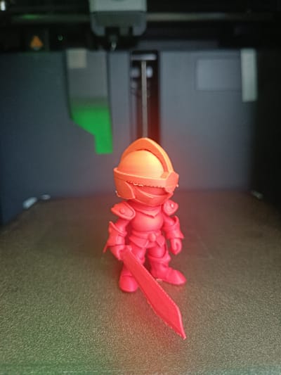 The image shows a 3D-printed miniature figurine of a knight, predominantly red and orange in color. The knight is standing upright, holding a sword in its right hand, which is positioned in front of its body. The figurine is placed on a textured, green surface, possibly the print bed of a 3D printer, as indicated by the blurry background which appears to be the interior of a 3D printer with some of its mechanical components visible, including a green light. The lighting in the scene is dim, casting a warm glow on the figurine. There is no visible text in the image. The context provided suggests the image was taken in Vratsa, Bulgaria.