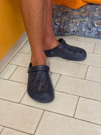 The image shows a close-up view of a person's legs and feet wearing navy blue Crocs. The person is standing on a tiled floor, which appears to be in a residential or commercial indoor setting. The tiled floor is light beige with darker grout lines. To the left of the legs, there is a wall painted in a bright yellow color. In the background, there is a bed covered with a blue blanket that has a pattern of cartoon cats and paw prints. An orange textile is visible above the blanket. The lighting suggests an indoor environment, possibly during the day. The image does not provide any specific indicators of its location within San Benedetto del Tronto, Italy.