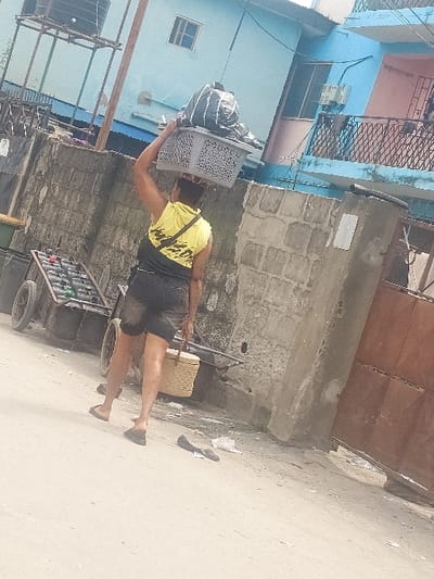 The image depicts a street scene in Agani, Nigeria. A person, viewed from the back, walks on an unpaved or lightly paved surface. This individual wears a yellow sleeveless top with a black graphic, dark shorts, and dark flip-flops. They carry a grey plastic laundry basket on their head, which contains a dark, bulky, possibly fabric-wrapped item. A light-colored woven basket is held in their right hand. To the left, a two-wheeled metal pushcart is visible, loaded with multiple dark-colored objects, possibly bottles or containers. A pair of dark flip-flops lies on the ground to the right of the person's path. In the background, a low concrete block wall extends horizontally. Behind the wall, several buildings are visible, including a prominent light blue multi-story structure with a small square window on the upper floor and a metal frame supporting a water tank on its roof. To the right of the blue building, another section features a pink-colored wall, a balcony with black metal railings, and two outdoor air conditioning units. A large, rusty brown metal gate is partially visible on the far right, adjacent to the wall. Scattered debris is present on the ground.
