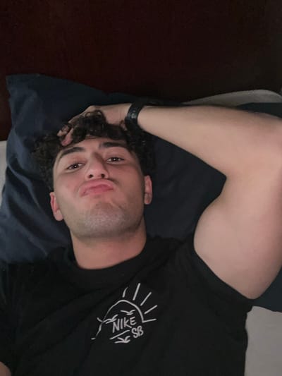 A single adult male is depicted in a close-up, high-angle shot, lying supine in a dimly lit indoor setting. He possesses dark, curly hair and light facial stubble. His lips are pursed, and he gazes directly towards the camera. He is wearing a black short-sleeved t-shirt featuring a white graphic design on the left chest area, which includes a sun, birds, and the text "NIKE SB." A dark-colored watch or wrist device is visible on his left wrist. His left arm is bent, with his hand resting on the back of his head, exhibiting visible bicep musculature. He is positioned on a dark blue pillow and dark bedding, with a dark, possibly wooden, headboard partially visible in the upper background. The environment is consistent with a bedroom. The image contains no identifiable visual cues to confirm the requested location of Sarouja Municipality, Syria.