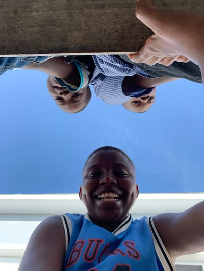 This image, taken from a low-angle perspective in Ibasa, Nigeria, features three individuals looking downwards against a clear blue sky. On the left side, a hand with light brown skin grips a wooden beam. Below this hand, a person with dark skin, wearing a striped shirt and dark pants, looks down. Further below and to the left, a second person, a child with dark skin and a striped shirt with blue trim, also gazes downwards. On the right side of the frame, a third individual with dark skin smiles broadly, showing their teeth. This person wears a bright blue jersey with the word "BULLS" partially visible in reverse text on the fabric. The background is dominated by a bright, cloudless blue sky, with a white vertical structure visible on the far right edge of the frame. The overall impression is one of a cheerful and engaging interaction, with the subjects appearing happy and curious as they look down towards the camera on a sunny day.