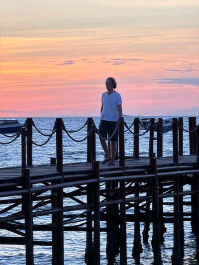 A person stands on a pier at sunset. The sky is a gradient of soft oranges and pinks. The person is wearing a white t-shirt and dark shorts. They are facing away from the camera, looking out at the water. The pier is made of dark wood and has a railing. The water is calm and reflects the colors of the sky. In the distance, there appears to be a boat. The scene evokes a sense of peace and tranquility. The time of day is sunset, and the weather is clear. The location is Baan Bang Rak, Thailand.