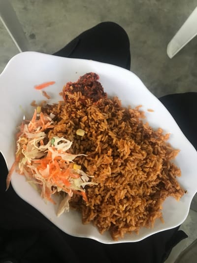 A white plate is filled with cooked rice, which is brown. Next to the rice is a piece of fried chicken, which has a darker brown color. A side of coleslaw is also present on the plate, consisting of shredded cabbage and carrots. The plate is set on a surface that includes black fabric.
