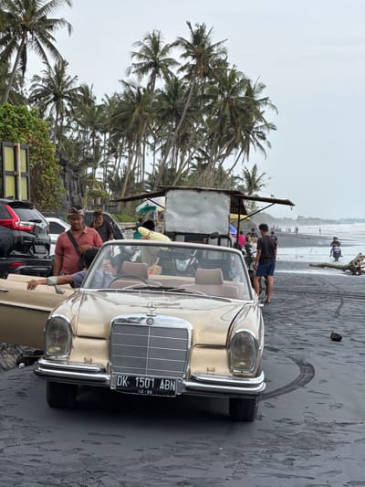 The image displays a black sand beach in Beraban, Indonesia, under an overcast sky. In the foreground, a vintage, light gold Mercedes-Benz convertible is parked on the dark sand. It bears an Indonesian license plate, "DK 1501 ABN," with "12-26" printed below it. The driver's side door is open, revealing tan interior seats.
To the left of the vehicle, a man wearing a reddish-brown short-sleeved shirt and a traditional Balinese head covering (udeng) stands, looking towards the viewer. His left arm rests on the open car door. Partially visible inside the driver's seat is a person wearing glasses. Behind the standing man, another man with a similar head covering is partially obscured. A black Honda CR-V and other vehicles are parked behind the Mercedes, near a low stone structure.
On the right, a rudimentary vendor cart with a light-colored surfboard attached to its rear is present. A man wearing a dark t-shirt and shorts stands next to a white scooter near the cart, facing the ocean. The background features numerous tall palm trees and the ocean, where several individuals are visible along the shoreline and in the shallow water. One person is operating a scooter in the water. Tire tracks are distinctly visible on the black sand in front of and to the right of the Mercedes. A piece of driftwood lies near the water's edge in the mid-ground.