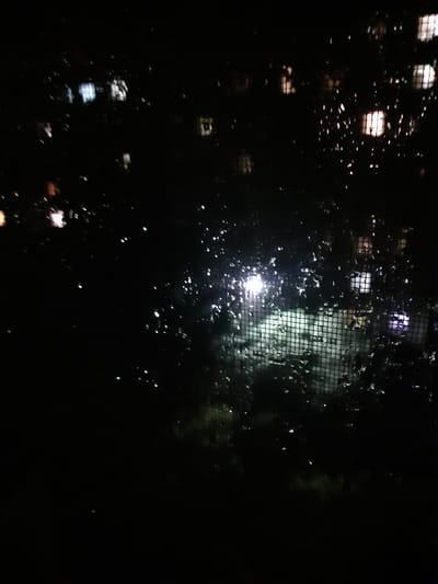 The image depicts a nighttime view through a window with raindrops on the glass, obscuring the scene outside. In the background, lights from apartment buildings or windows are visible as blurry, rectangular shapes. Some lights appear white or yellowish, suggesting interior lighting, while others have a purplish hue, possibly indicating exterior lights or reflections. A faint grid pattern, likely from a screen or blinds, is visible in the middle ground. The overall environment appears to be urban, consistent with a city like Obukhiv, Ukraine. No people or specific actions are discernible due to the low light and the water droplets on the window. The scene conveys a sense of looking out into a dark, wet night.