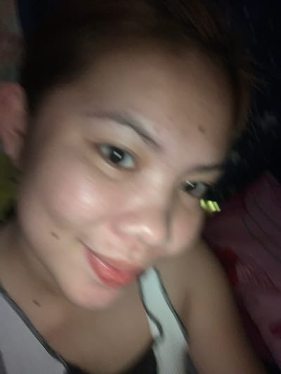 A selfie is taken from a close-up, low-angle perspective of a young woman with light brown skin and dark brown hair. Her face is slightly blurred, but she appears to be smiling with her mouth slightly open, revealing a glimpse of her lips. She is wearing a dark-colored top with white trim. The background is dark and appears to be indoors, with some blurry hints of bedding or fabric. There is no discernible indication of the city of Kiblawan, Philippines.