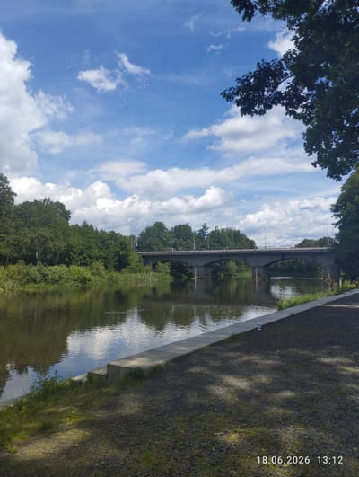 A wide, calm river flows towards the viewer, reflecting the sky and surrounding greenery. On the far bank, dense trees line the water's edge, partially obscuring a stone bridge with multiple arched spans. Several streetlights are visible on the bridge. On the near bank, a paved area with gravel and sparse grass leads down to a concrete embankment and steps. Shadows of tree branches are cast across the paved area. The sky is blue with scattered white clouds. A date and time stamp, "18.06.2026 13:12," is visible in the lower right corner.