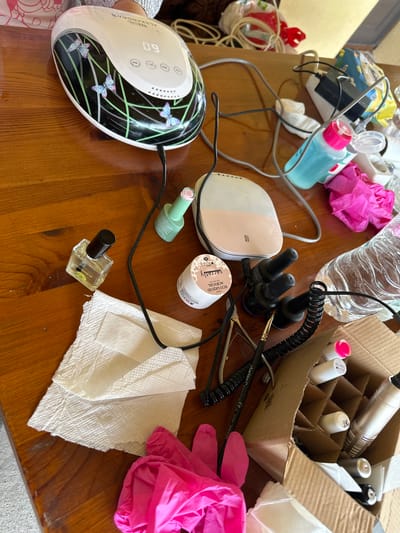 The image shows a wooden table surface cluttered with various nail care items. A nail lamp with butterfly decorations sits on the left side of the frame. Nearby are bottles of nail polish, including one clear bottle labeled "SNB nourishing oil". There's also a container labeled "ECO LIQUID ACRYGEL COLLECTION" from "secretly". A cardboard box holding nail polish bottles is on the right. Several pink gloves, white paper towels, and a water bottle are scattered around the table. A brush labeled "clarina Connello CL7319 Made in Italy" is also visible. The location in the city of Lehchevo, Bulgaria is not discernible.